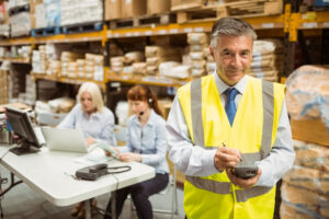 A male business owner in his warehouse wearing hi-vis with staff members in the background