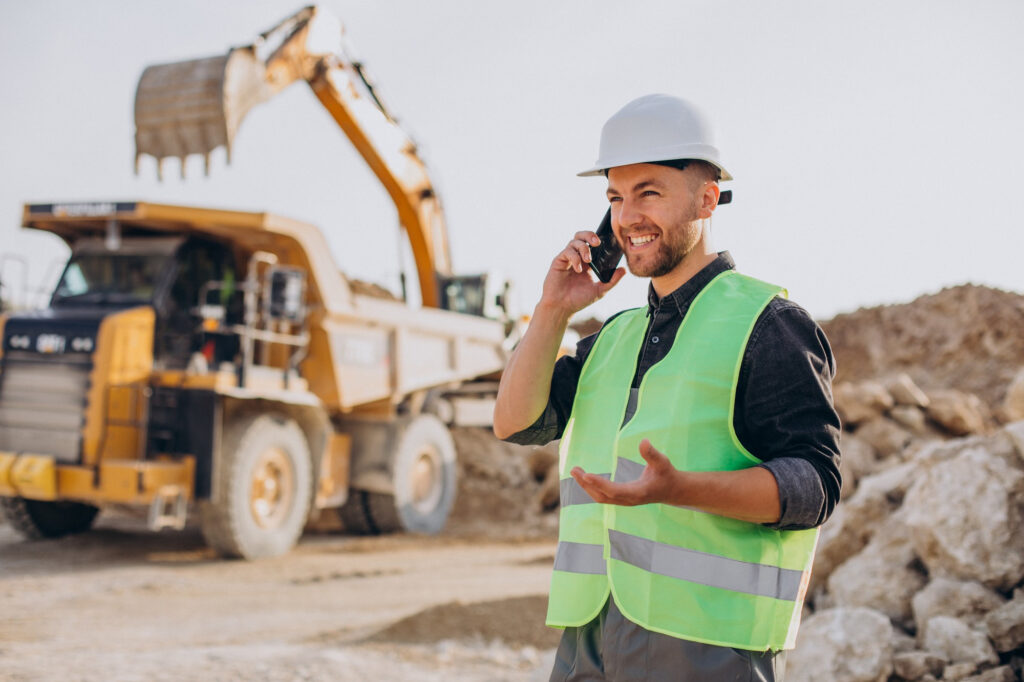 A worker wearing high vis on the phone with large truck and excavator in the background